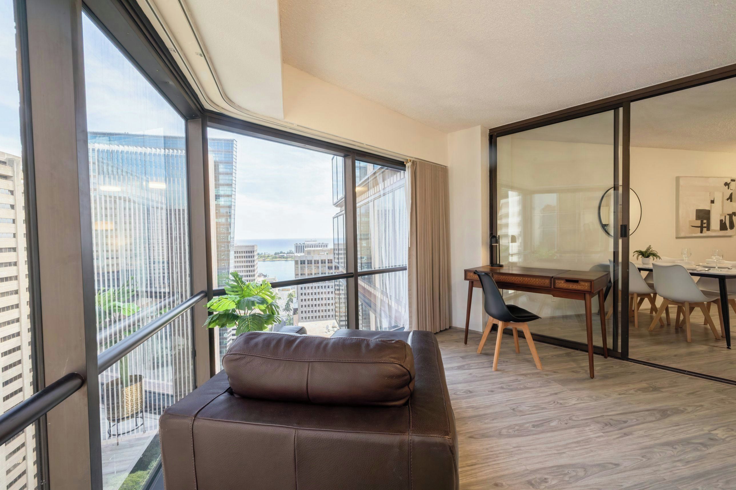A modern living room with a large window, a sofa, a dining area with a table and chairs, and a view of the cityscape.