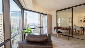 A modern living room with a large window, a sofa, a dining area with a table and chairs, and a view of the cityscape.