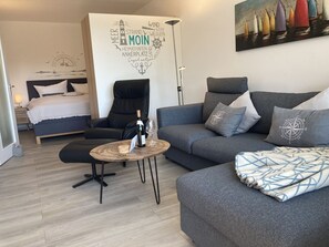 A modern living room with a bed, a black leather chair, a grey sofa, and a round coffee table. There is a wall with nautical-themed decor and a painting of colorful boats.