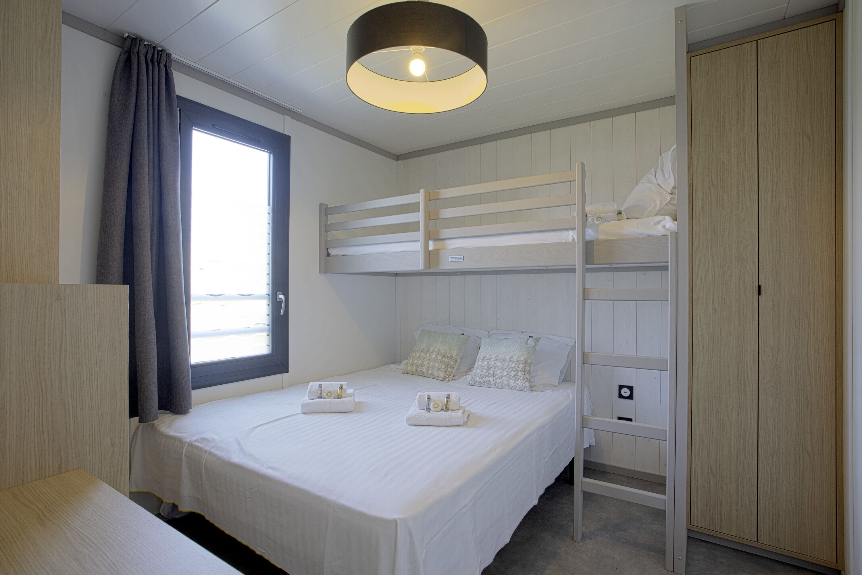 A bunk bed room with a window, a bedside table, and a wardrobe.