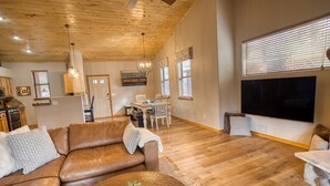 A spacious living room with a brown leather sofa, a round coffee table with a plant, a dining area with a table and chairs, wooden flooring, and a large TV on the wall. - Torreon Cardinal Landing Cabin With Party Garage (Show Low)