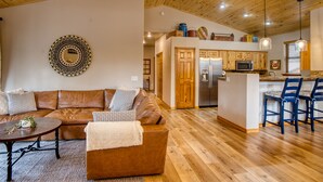 A spacious living area with a brown leather sofa, a coffee table, and a patterned rug. A kitchen with wooden cabinets and a stainless steel refrigerator is visible in the background. - Torreon Cardinal Landing Cabin With Party Garage (Show Low)