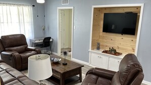 A living room with a brown leather sofa, a wooden coffee table, a TV mounted on a wooden wall, and a ceiling fan.