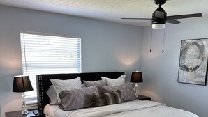 A bedroom with a bed, two bedside tables, a ceiling fan, a window with blinds, and a wall-mounted abstract painting.