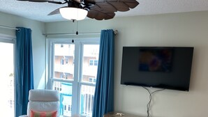 A living room with a ceiling fan, a white armchair, a patterned sofa, a small round table, and a flat-screen TV mounted on the wall.