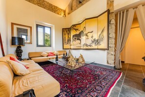 A living room with a sofa, a glass coffee table, a red patterned rug, a painting, and a stone wall.