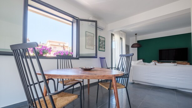 A modern living room with a large window, a dining table with chairs, a sofa, and a flat-screen TV.
