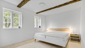 A bedroom with a wooden floor, a white bed, two windows, a bedside table with a lamp, and exposed wooden beams on the ceiling. - Amazing apartment in Tisvildeleje (Tisvildeleje)