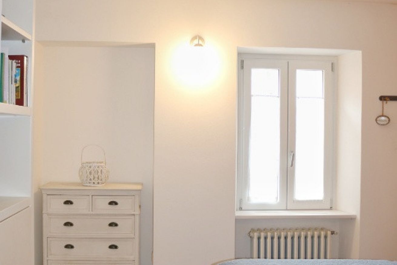 A bedroom with a white dresser, a window with blinds, a radiator, and a wall-mounted light fixture.