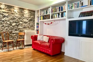 A living room with a red sofa, wooden chairs, a bookshelf, a stone wall, and a television. - My Heart in Argegno - My Home in Como (Argegno)