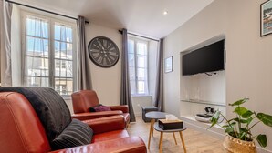 Un salon moderne avec un canapé en cuir, une petite table, une plante en pot, un téléviseur fixé au mur et une grande horloge murale.