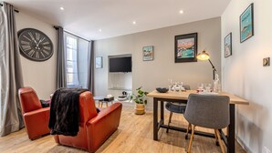 Un salon moderne doté d’une grande horloge murale, d’un téléviseur à écran plat, d’une table à manger avec des chaises et d’une grande fenêtre agrémentée de rideaux.