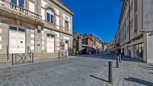 Une rue pavée bordée de bâtiments historiques, une entrée fermée par un portail et un ciel d’un bleu éclatant.
