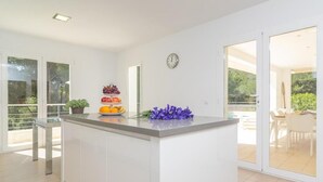 Une cuisine moderne avec un îlot central, des meubles blancs et une horloge au mur, donnant sur la terrasse extérieure.