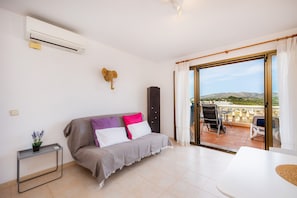 Une pièce comprenant un canapé, une petite table avec une plante, un climatiseur, un meuble et une porte-fen être coulissante donnant sur un espace extérieur avec une table et des chaises.