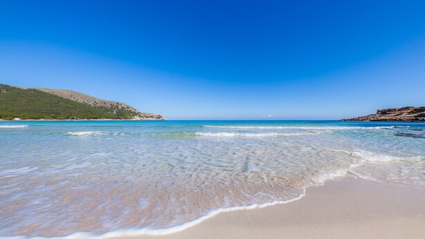 Een strand met helder water, een zandstrand en verafgelegen rotsheuvels.
