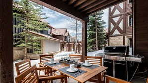 Une terrasse extérieure avec des tables et des chaises en bois, un barbecue et une vue sur un bâtiment en bois doté de fondations en pierre.