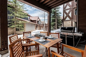 An outdoor patio with wooden tables and chairs, a grill, and a view of a wooden building with a stone foundation.