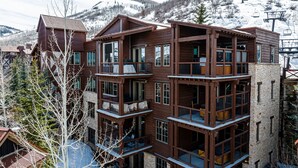 Un immeuble en bois à plusieurs étages, doté de balcons et d’une façade en pierre, avec un télésiège visible en arrière-plan.
