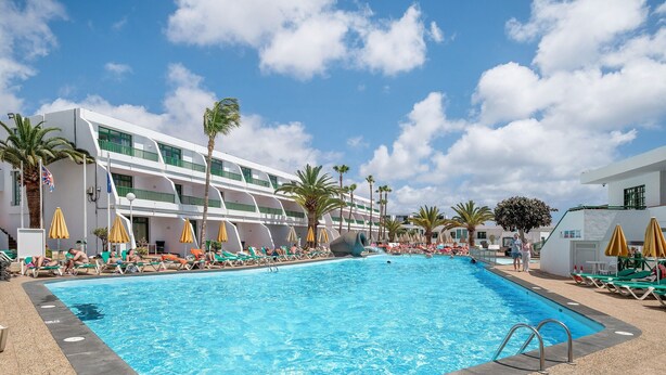 Una piscina con sillones de descanso y sombrillas, rodeada de palmeras y un edificio moderno con balcones.