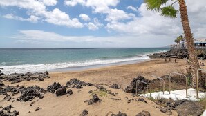 Una playa con terreno rocoso, una palmera y un edificio con techo azul.