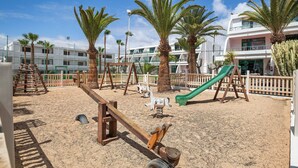 Un parque infantil de arena con columpios, un tobogán y palmeras.