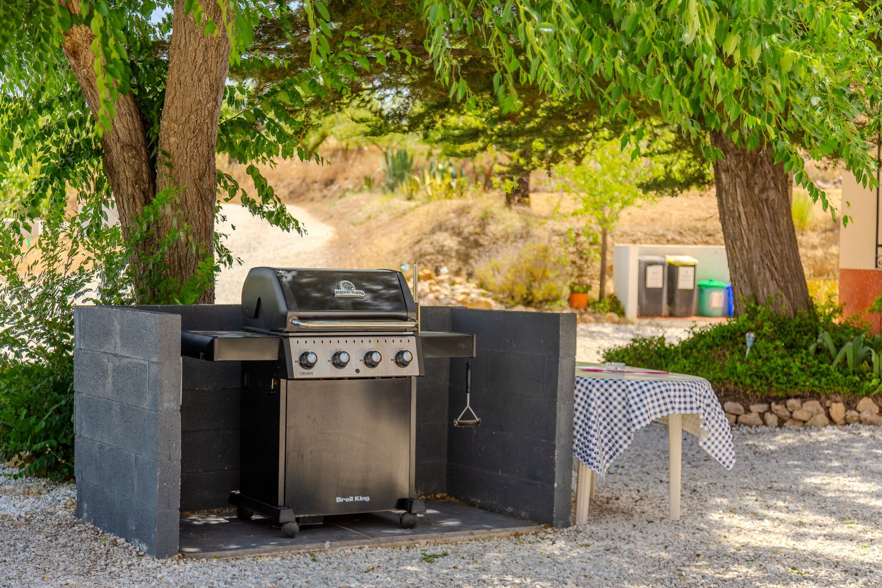 Una parrilla portátil con cuatro quemadores, una mesa con mantel a cuadros y un suelo de grava.