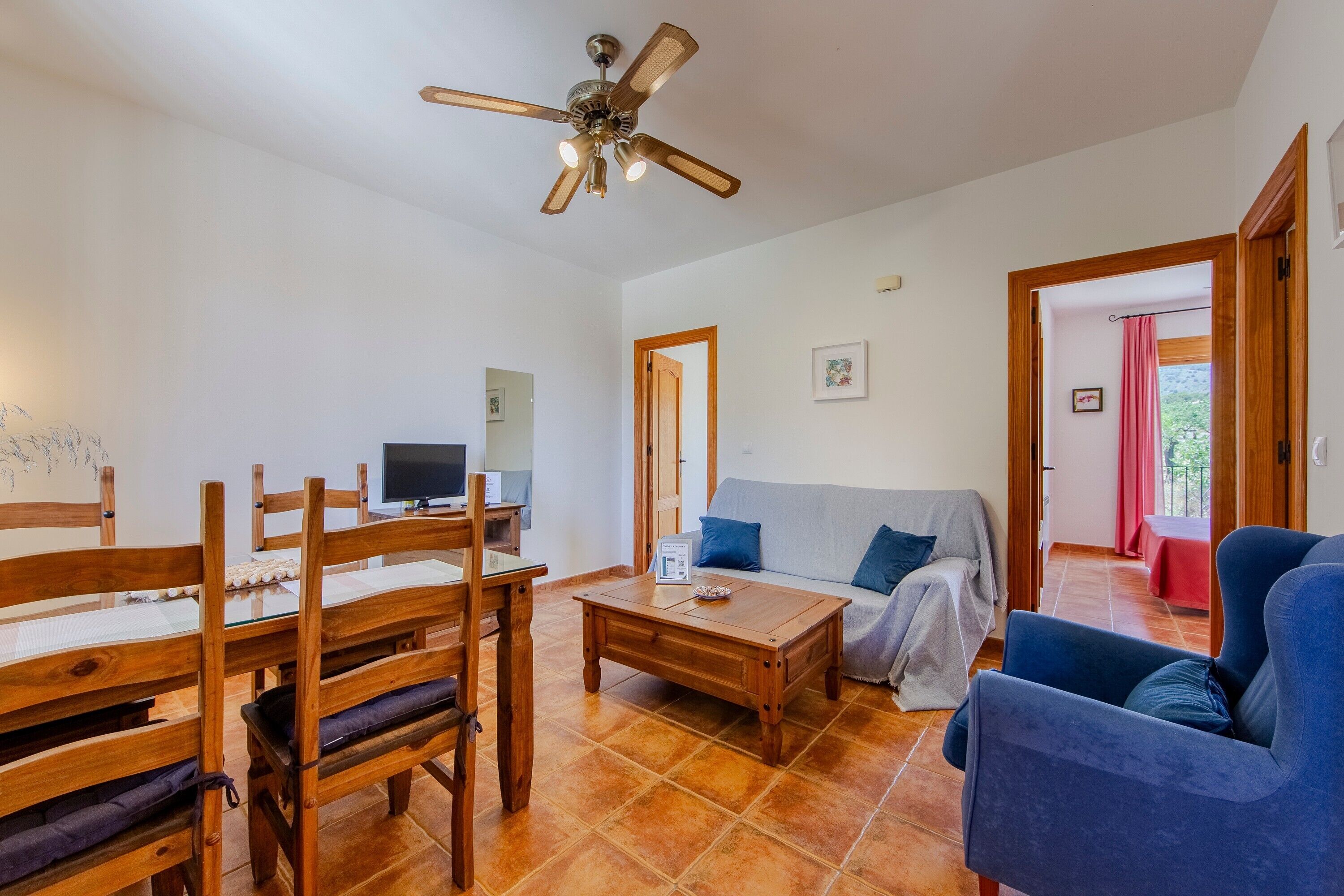 Una sala de estar con una mesa de comedor de madera, sillas, un sofá, una mesa de centro, un ventilador de techo y un televisor.