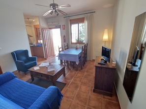 Een woonkamer met een bank, een eettafel met stoelen, een televisie op een standaard en een plafondventilator.