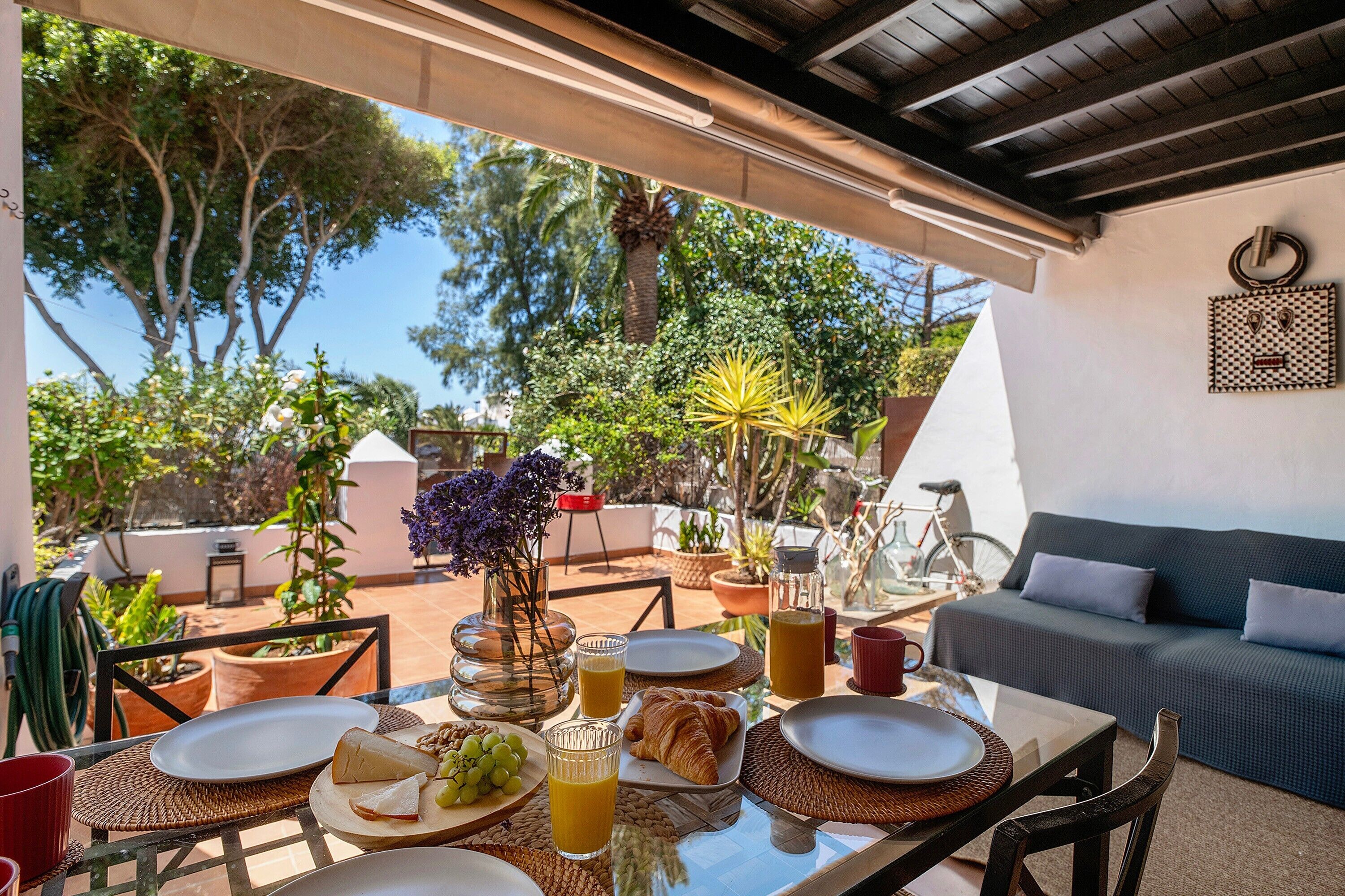 An outdoor dining area with a table set for breakfast, surrounded by potted plants and a view of trees.