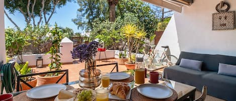 An outdoor dining area with a table set for breakfast, surrounded by potted plants and a view of trees.