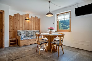 Um quarto acolhedor com um banco de madeira, uma mesa redonda de madeira com cadeiras, uma televisão fixada na parede e uma janela com vista para as montanhas.
