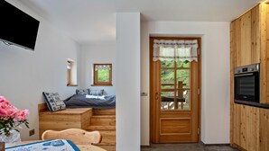 Eine kleine Küche mit Einbauofen, einem Holztisch mit gemustertem Tischläufer, einer kleinen Holzbank und einem Bett mit blau-weißem Kopfkissen.