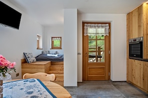 A compact kitchen with a built-in oven, a wooden dining table with a patterned tablecloth, a small wooden bench, and a bed with a blue and white pillow. - Apartment 'Salto Del Camoscio' with Mountain Views and Private Garden (Rabbi)