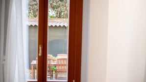 Vista a través de una puerta de madera con ventana, mostrando un área de estar al aire libre con mobiliario de madera y una pequeña mesa.
