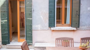 Una habitación con una mesa y sillas de madera, un ventanal con contraventanas verdes y una puerta que da a otra estancia.