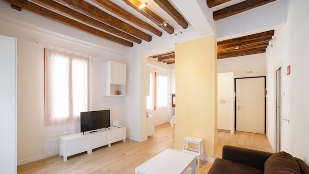 Un salón moderno con televisor, muebles blancos, vigas de madera y un pasillo que conduce a una puerta.
