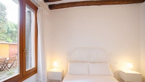 Un dormitorio con cama, dos mesitas de noche con lámparas, una ventana con cortinas y vistas a un espacio exterior con mesa y sillas.