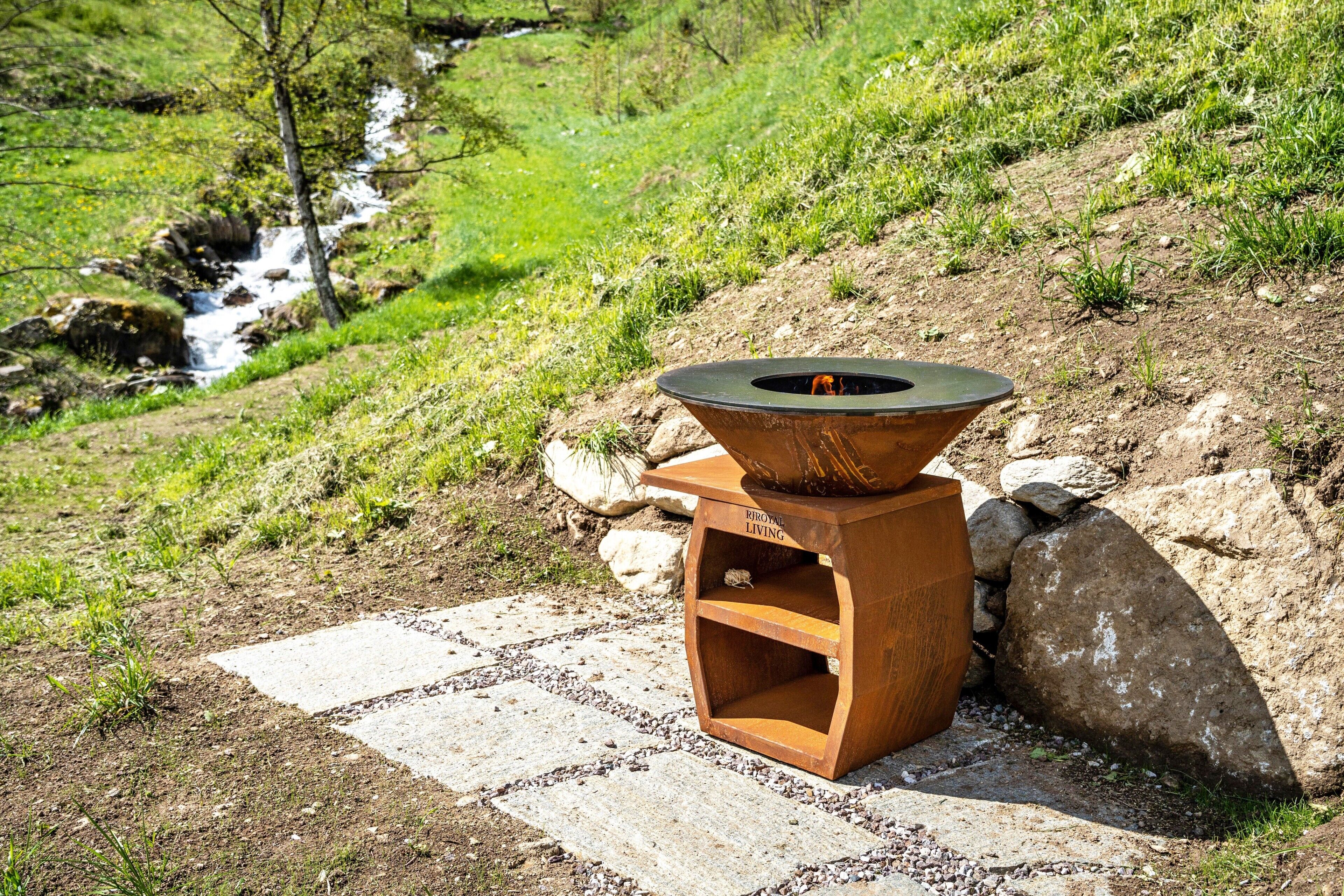 A rustic outdoor fire pit with a metal bowl, surrounded by a natural landscape with a stream and greenery.