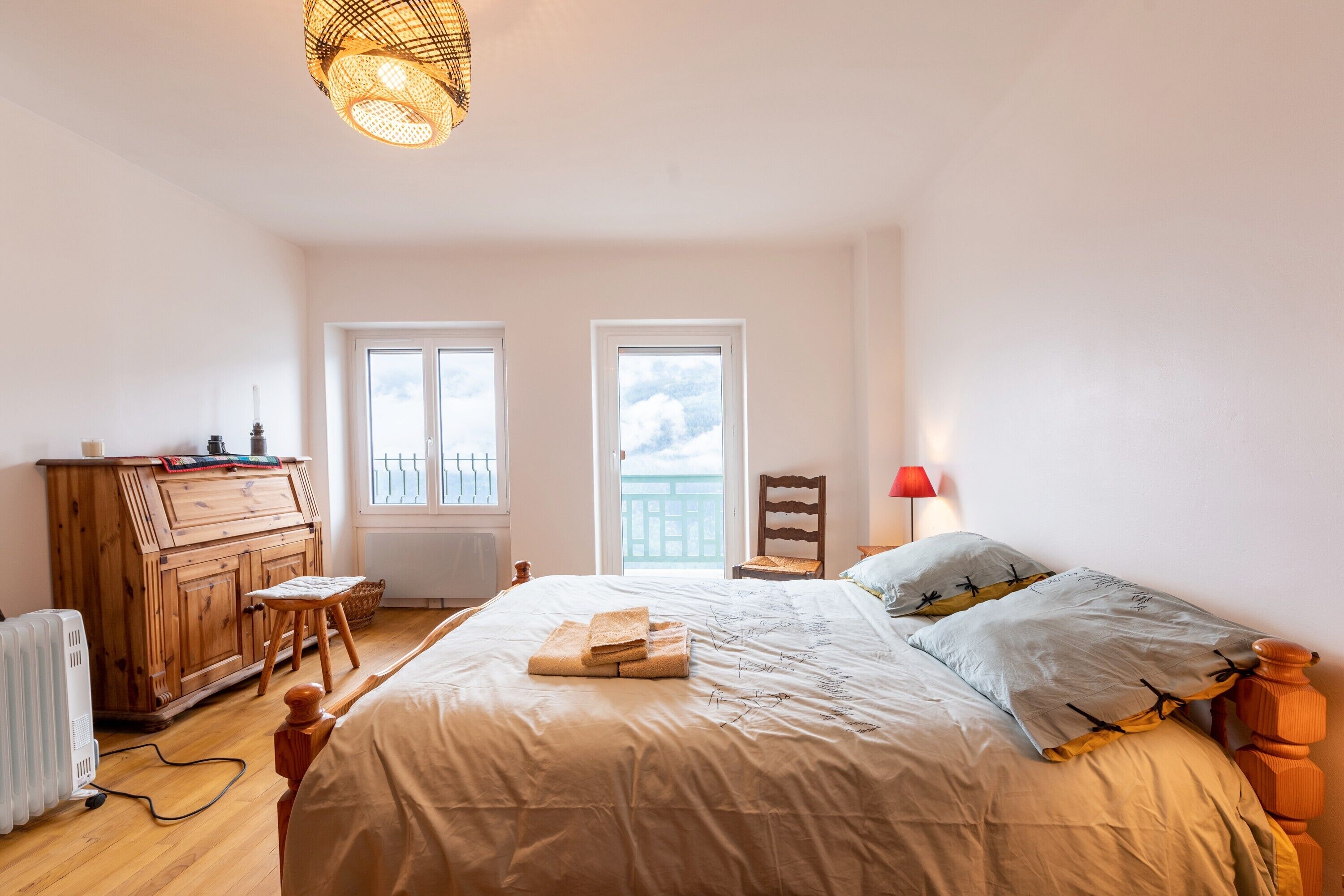 A bedroom with a bed, wooden dresser, chair, radiator, and a view of the sky through two windows.
