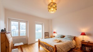 Une chambre avec un lit en bois, deux fenêtres, un radiateur, une commode en bois, une chaise, une table de chevet avec une lampe et une bibliothèque.