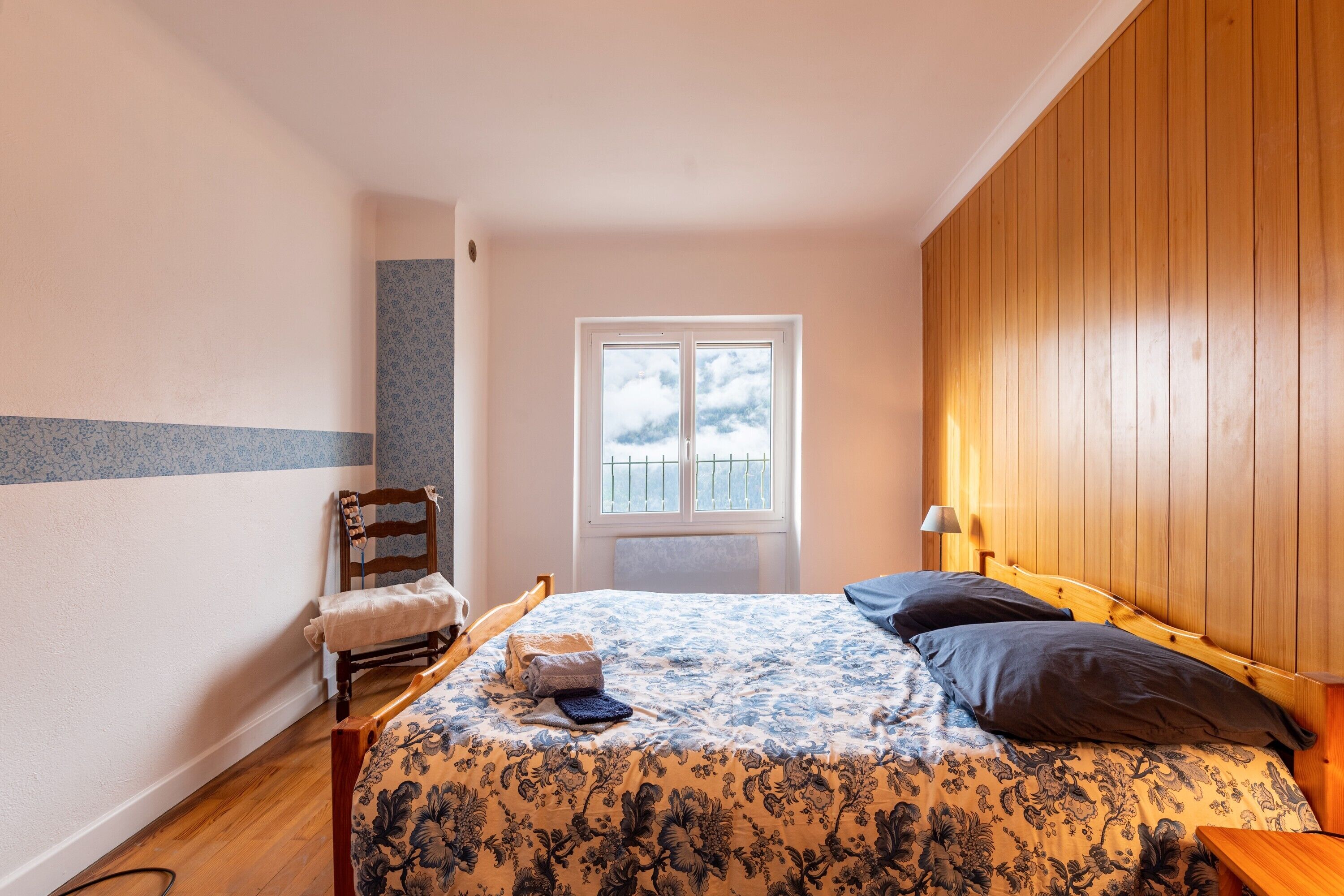 Une chambre à coucher avec une tête de lit en bois, un lit recouvert d’une literie à motifs floraux, une chaise, une fenêtre et une lampe.