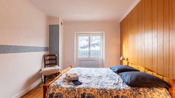Une chambre à coucher avec une tête de lit en bois, un lit recouvert d’une literie à motifs floraux, une chaise, une fenêtre et une lampe.