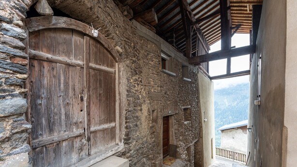 Un bâtiment en pierre doté de portes en bois et d’un escalier menant à un balcon offrant une vue sur la montagne.