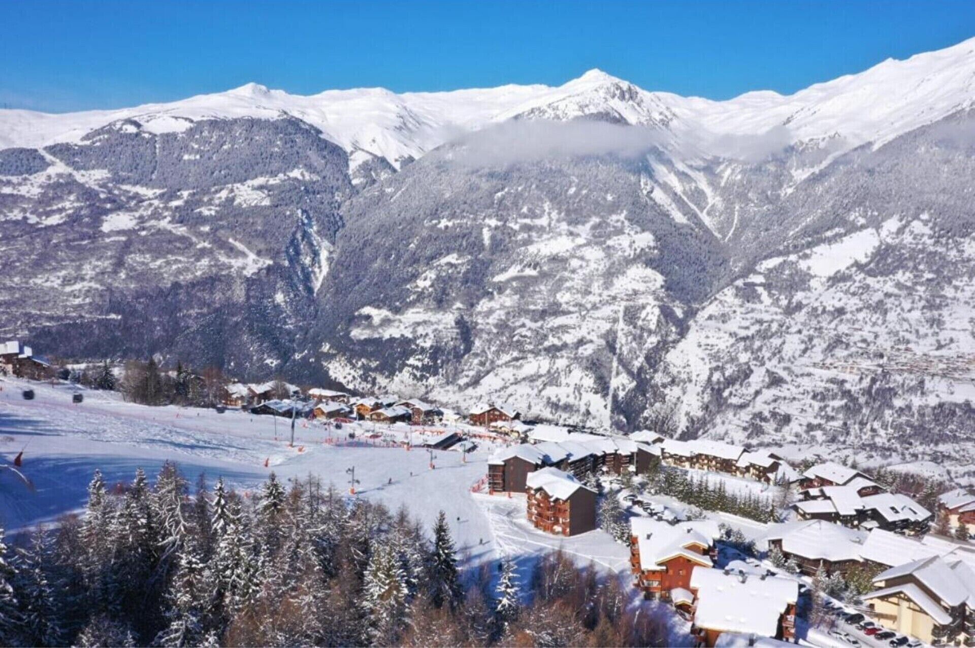 Un village de montagne enneigé, avec ses bâtiments traditionnels en bois, une piste de ski et un ciel bleu azur.