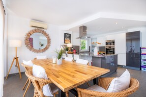 Cozinha moderna com eletrodomésticos de aço inoxidável, área de jantar com mesa de madeira e cadeiras de vime, e um espelho grande na parede.