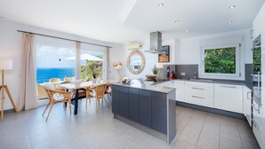 A modern kitchen with a large island, dining area with wicker chairs, and a view of the ocean. - Villa 'Azul' with Shared Pool, Wi-Fi and Air Conditioning (Cala Pi)