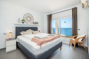 Quarto com cama, mesa de cabeceira, abajur, relógio na parede e vista para o mar.