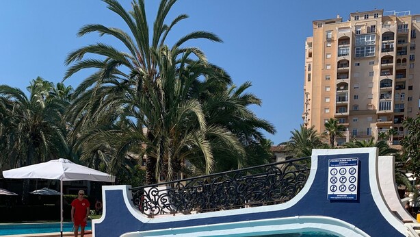 Una piscina con un arco decorativo, palmeras y un edificio al fondo.