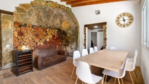Ein Kamin aus Stein mit Holzscheiten, ein braunes Ledersofa, ein Holzesstisch mit weißen Stühlen und eine Wanduhr.